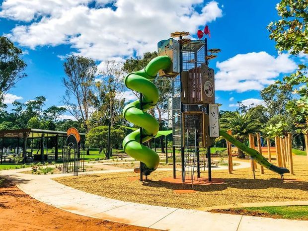 A bright outdoor playground in a landscaped park, featuring a tall multi-level climbing structure with a prominent green spiral tube slide, climbing nets, platforms, and a smaller straight slide. Paved paths, play surfacing, shade structures, trees, and lawn surround the equipment under a vivid blue sky.
