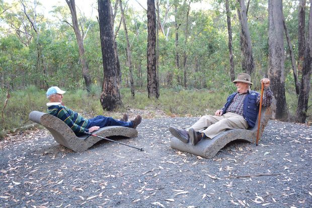Two people are reclining on sculptural curved outdoor seats along a gravel path in a eucalyptus woodland. The setting is a natural bushland landscape, with textured concrete or stone-like seating integrated into the outdoor environment.