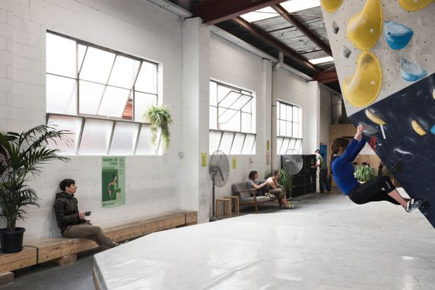 Interior view of a spacious industrial-style indoor climbing gym. The high-ceilinged space has white painted masonry walls, exposed dark steel roof framing, large metal-framed windows, timber bench seating, industrial fans, and a large climbing wall with colourful holds at right. Several people are climbing or resting, with potted plants adding greenery.