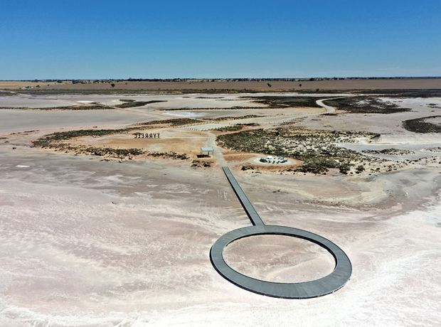 Aerial view of a stark, flat salt-lake or salt-pan landscape with pale pink, white and beige mineral surfaces. A narrow dark boardwalk extends from the middle distance to a large circular viewing platform in the foreground, with sparse low vegetation and exposed salt formations scattered across the terrain.