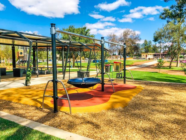 A bright outdoor playground in a landscaped park, featuring a modern metal-framed shade structure, suspended circular swing equipment, colourful red and yellow safety surfacing, picnic seating, paved paths, lawns and mature trees under a blue sky.