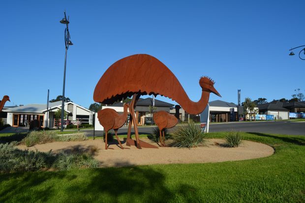 A landscaped public streetscape featuring a large rust-coloured metal emu sculpture with smaller emu figures, set in a sandy garden bed with grasses and lawn. Low-rise commercial or civic buildings and distinctive street lighting are visible in the background beneath a clear blue sky.