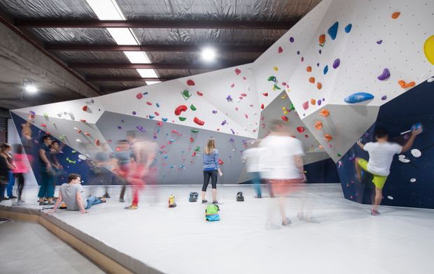 Indoor bouldering and climbing gym with angular white climbing walls covered in colourful holds, a padded light-grey floor, exposed industrial roof structure, and several climbers and visitors. The space is brightly lit and energetic, with dark blue wall sections creating contrast.