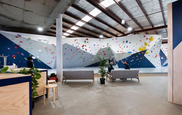 Interior view of a modern indoor climbing facility with extensive geometric climbing walls covered in colourful holds. A polished concrete floor, exposed insulated roof structure, white cylindrical columns, lounge seating, plants, and a small reception or service counter are visible. The space is bright, open, and energetic, with industrial architectural elements and a playful climbing-focused fit-out.