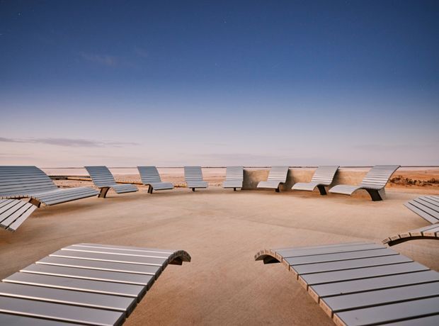 An expansive outdoor terrace or poolside-style area with rows of sculptural silver-grey slatted chaise lounges arranged across a pale sandy surface. The space opens toward a flat, arid horizon beneath a dramatic blue-to-pink sky, creating a minimalist and surreal desert-like setting.