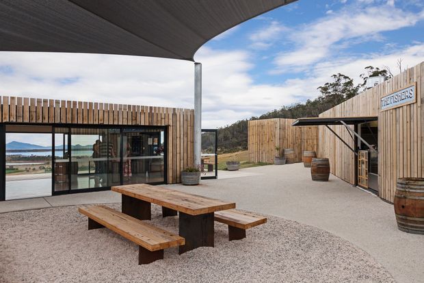 A landscaped outdoor courtyard or terrace beside a contemporary timber-clad building, viewed beneath a broad curved roof canopy. The space features a solid timber picnic table, gravel and concrete paving, timber privacy fencing, planting pots, and barrel-style decorative elements. Glazed sliding doors reveal an interior space, while a distant water and hill landscape is visible beyond the building.