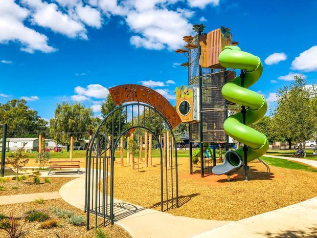 A bright outdoor playground in a landscaped park, featuring a tall multi-level timber and metal play structure, a prominent lime-green spiral tube slide, climbing elements, and an arched metal entry feature. The playground sits on a tan sand or soft-fall surface with curved concrete paths, benches, low planting, and mature trees under a vivid blue sky.