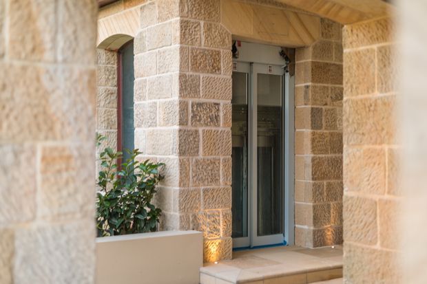 Close view of a refined stone-clad entryway framed by substantial sandstone masonry piers and an arched opening. A glazed contemporary door sits above tiled steps, with recessed warm lighting at the base of the walls and a built-in planter containing greenery. The image combines traditional masonry detailing with a modern glazed entrance.