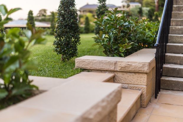Close-up view of landscaped residential entry steps and low retaining walls, finished in warm beige textured masonry with matching paved surfaces. Black metal stair railings border the steps, while manicured lawn, clipped shrubs, and ornamental planting surround the entry.