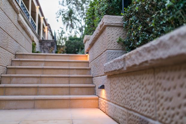 A landscaped exterior stairway with broad pale stone or porcelain-tiled steps ascending between textured masonry retaining walls. Integrated warm lighting illuminates the side of the stair, while dense hedging and trees frame the path. A building facade is partially visible along the left edge.