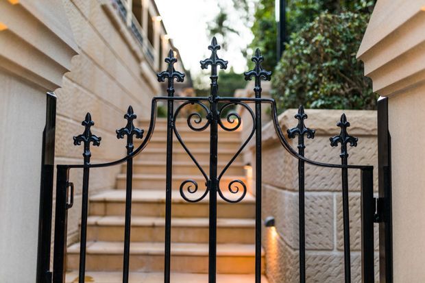 Close-up view of an ornate black wrought-iron pedestrian gate with decorative spearhead finials and curled scrollwork, opening onto a narrow tiled stairway. The gate is framed by pale rendered and textured masonry walls, with warm step lighting and greenery beyond.
