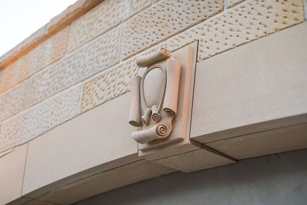 Close-up architectural detail of a pale stone or concrete facade featuring a projecting sculptural ornamental bracket with curled scroll motifs. Above it is a band of perforated or textured masonry blocks, with a warm timber-toned trim along the upper edge. The image focuses on the building envelope and decorative craftsmanship rather than an interior space.