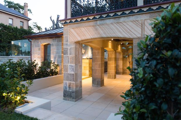 A refined sandstone-clad residential courtyard or entry pavilion, viewed from an outdoor garden area. The covered structure features substantial stone columns, large stone floor tiles, warm recessed lighting, an ornate dark metal balustrade above, and a decorative scalloped roof-edge detail. Landscaped hedges and raised garden beds frame the paved approach, while a glazed opening and illuminated interior are visible beyond the arcade. A neighbouring traditional brick residence is visible in the background.