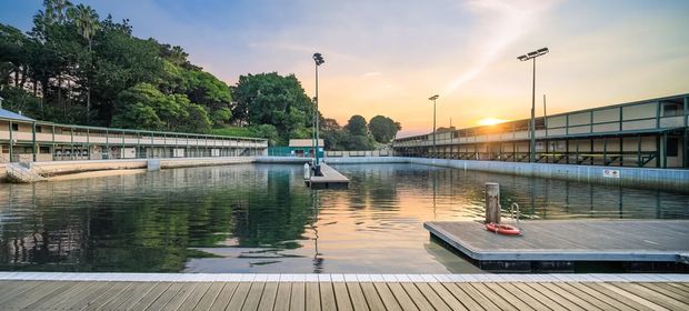 Wide outdoor view of a calm waterfront rowing or watersports facility at sunset, with floating timber docks in the foreground, a large rectangular basin, long low spectator or training buildings, floodlights, and dense tropical landscaping beyond.