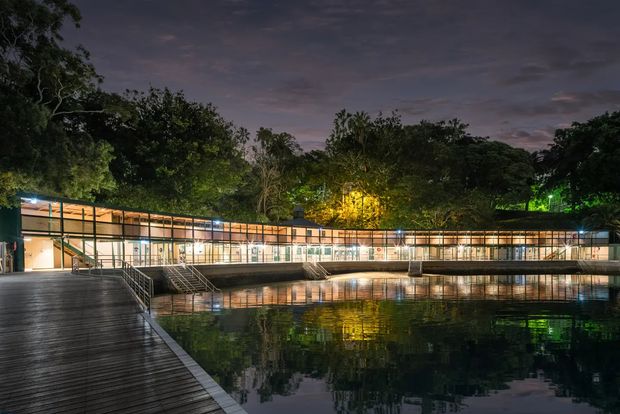 A long, low modern building is illuminated at dusk beside a still reflecting water body. Extensive glazed walls, warm interior lighting, slender dark structural framing, and a broad timber boardwalk create a calm, resort-like setting surrounded by dense mature vegetation.