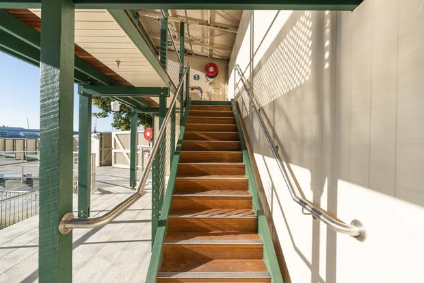 A bright exterior-access stairway on a raised or multi-level structure. The stairs have warm timber treads with green-painted metal or timber stringers and railings, alongside a pale wall with stainless-steel handrails. Open-sided circulation space, safety equipment, and strong patterned sunlight are visible, with an outdoor deck and distant urban or waterfront surroundings beyond.