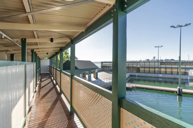 Covered elevated walkway with green-painted steel posts and beams, perforated metal balustrades, and a corrugated metal canopy. The walkway overlooks a large outdoor aquatic facility with visible pool structures and floodlights. Bright daylight and strong linear perspective create an airy, utilitarian circulation space.
