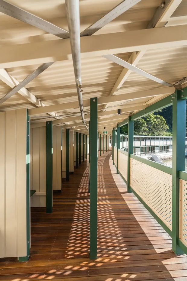 A long covered exterior walkway or verandah with dark timber decking, cream corrugated roofing and exposed structural bracing, green-painted posts and frames, and perforated cream balustrade panels. Repetitive columns and strong patterned sunlight create depth along the corridor, with a building and trees visible beyond the railing.