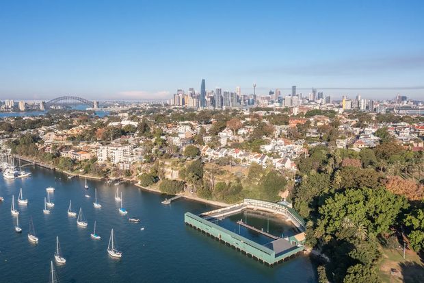 A wide aerial panoramic view of a Sydney harbour inlet with numerous moored sailboats, a waterfront swimming enclosure or pier in the foreground, dense leafy residential neighbourhoods, and the city skyline with high-rise towers and a large arched bridge in the distance. The image is dominated by the urban waterfront setting and landscape rather than a single building interior or facade.