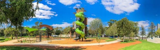 Panoramic outdoor view of a landscaped recreational playground, featuring a tall green spiral tube slide tower, climbing structures, shaded play areas, paved paths, lawn, mature trees, and bright blue sky.