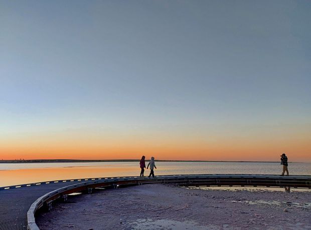 A wide coastal landscape at sunset, showing a curved boardwalk or viewing platform along a calm shoreline. Several people walk across the structure, with a broad orange-to-blue sky and low horizon in the background.