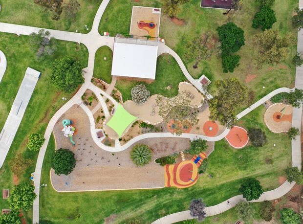 Aerial view of a landscaped outdoor recreational area with winding paved paths, expansive lawns, mature trees, landscaped garden beds, and multiple colourful playground and activity zones. Several covered structures and circular play areas are visible throughout the site.