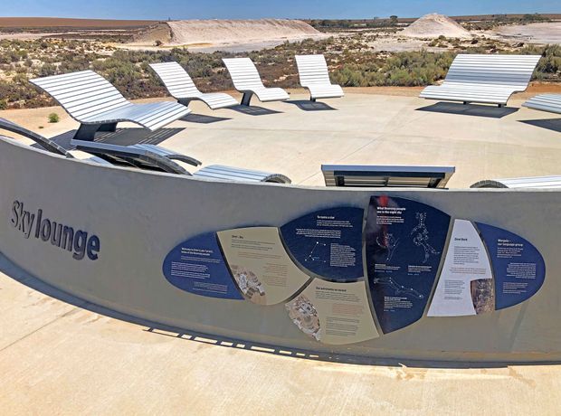 Outdoor interpretive exhibit labelled “SkyLounge,” featuring rows of curved silver lounge seats on a broad pale concrete platform in an arid, rocky landscape. An informational sign occupies the foreground, with low scrub and pale earth formations extending into the background.