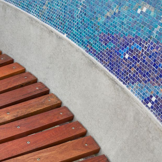Close-up detail of a swimming pool edge featuring reddish-brown timber decking beside a curved wall clad in small blue and turquoise mosaic tiles. The image emphasizes the contrast between warm wood, pale concrete, and vibrant patterned tile.