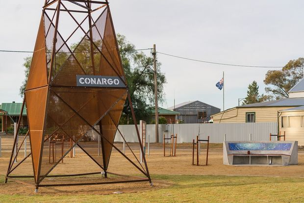 Exterior view of a rural or suburban community site featuring a tall rust-coloured steel geometric tower structure with a CONARGO sign, a flagpole, low corrugated-metal buildings, utility poles, young trees, and a concrete public seating or planter feature. The scene has an open, low-density character and appears to be a civic or community facility.