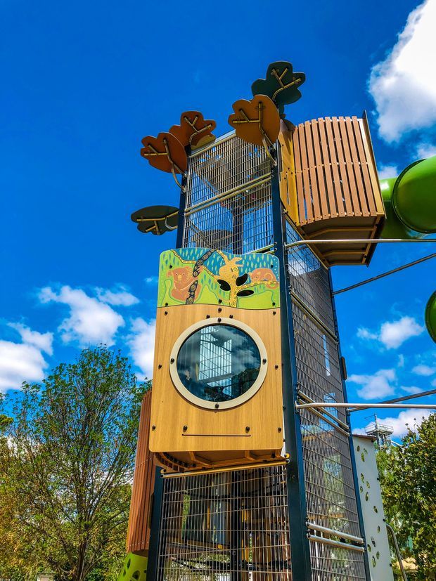 A tall, colourful outdoor children's play structure with a vertical metal climbing frame, timber-clad playhouse elements, circular window, decorative animal artwork, platforms, and green tube slides. The structure is photographed from below against a bright blue sky and trees.