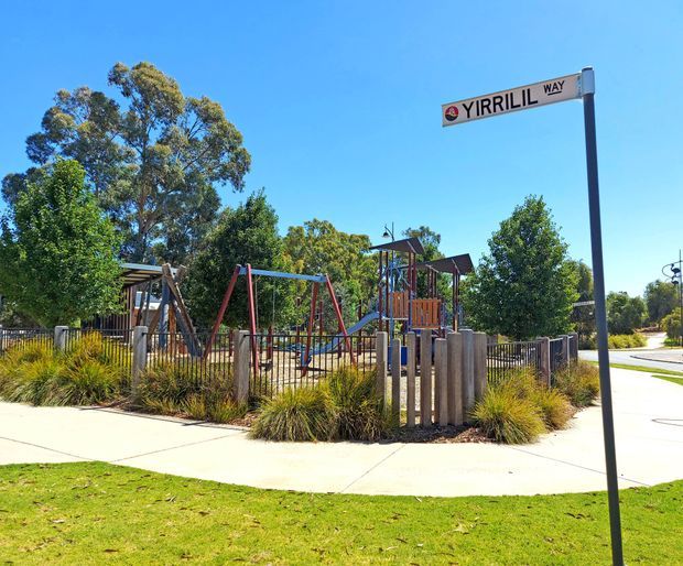 Bright daytime exterior view of a landscaped public playground beside a paved footpath, with swings, climbing structures, timber posts, metal fencing, ornamental grasses, mature trees, and a clearly visible Yirrill Way street sign. The scene has a contemporary suburban streetscape character.