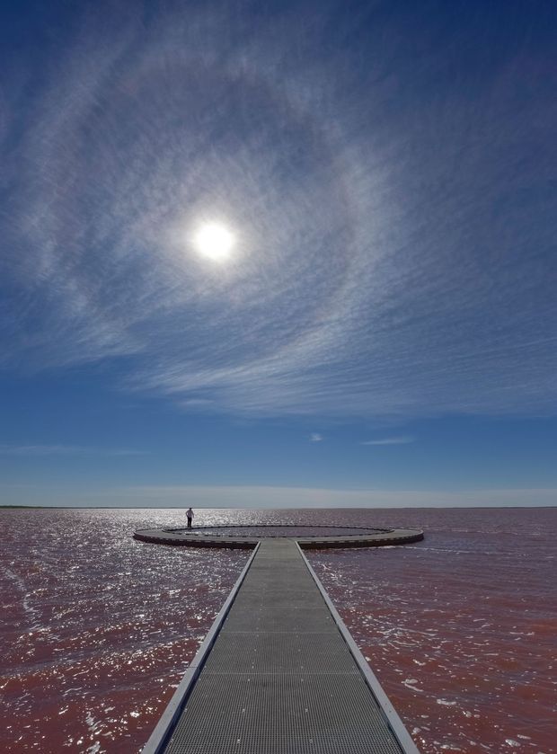 A long narrow grey walkway or pier extends across shimmering reddish water toward a circular viewing platform with a person standing on it. The image is dominated by a bright sun, expansive blue sky with wispy clouds, and a flat horizon, creating a strong geometric perspective toward the open water.