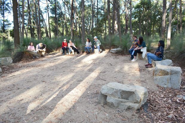 An outdoor gathering area in a wooded setting, with people seated around a broad sandy clearing on large rough-hewn stone benches and blocks. Tall trees, native grasses, leaf litter, and dappled sunlight create a natural bushland atmosphere.