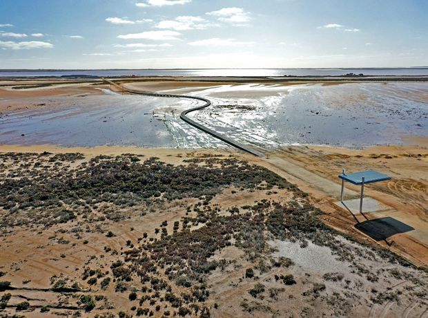 Aerial view of a broad coastal salt-pan or tidal wetland landscape, with shallow reflective water channels, sandy tracks, sparse low vegetation, and a small elevated blue-roofed shelter or structure in the foreground. The horizon and open water dominate the distant background.