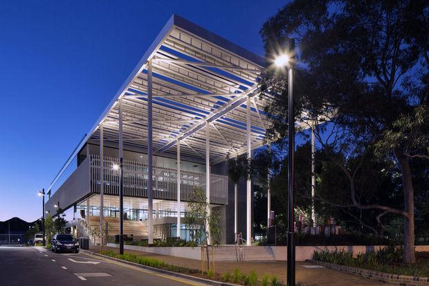 Dusk exterior view of a contemporary multi-storey building with a large illuminated open steel canopy, slender vertical structural supports, glazed ground-level frontage, and a landscaped street edge. Mature trees frame the right side, while street lighting and road markings emphasize an urban or institutional setting.