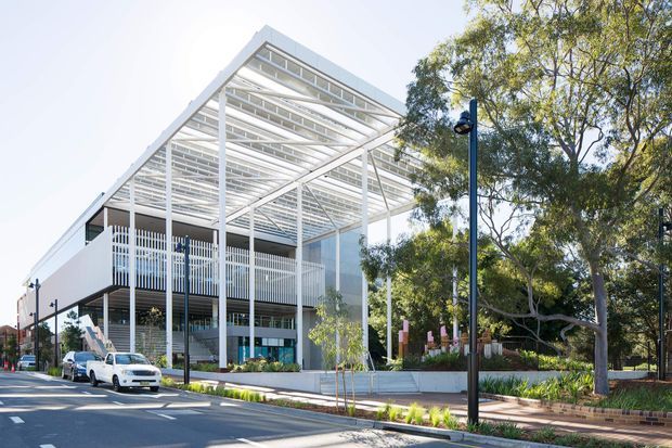 Street-level exterior view of a contemporary multi-storey building with a prominent white steel pergola or brise-soleil canopy extending over the facade and entry area. The building has extensive glazing, vertical screening elements, balconies, and a landscaped frontage in an urban setting.