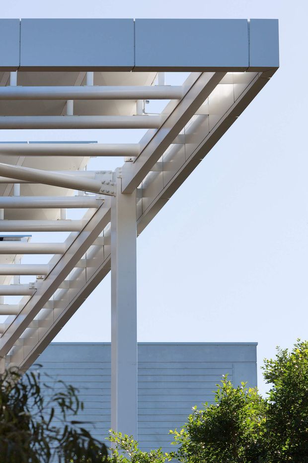 Close-up exterior architectural photograph looking upward at a modern structure with a projecting flat roof or canopy, exposed white-painted steel columns and tubular beams, and a blue-grey parapet or fascia. Horizontal metal cladding is visible on the building behind, with clear sky and landscaping at the edges. The image emphasizes the building's structural frame and cantilevered geometry.