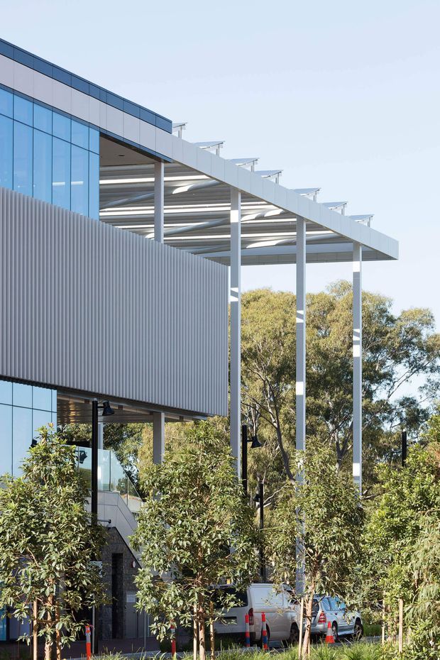 Exterior view of a contemporary multi-storey commercial building with extensive blue-tinted glazing, ribbed metal facade cladding, and a large projecting metal canopy supported by slender columns. Landscaping and trees partially screen the lower level, with service vehicles visible near the building.