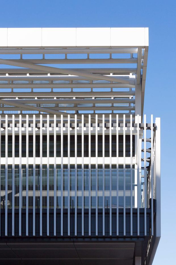Close-up upward view of a contemporary commercial building facade under a clear blue sky. The building features extensive glass curtain walling, white vertical sun-shading fins, dark metal lower panels, and a large exposed steel canopy or roof structure with horizontal beams and perforated framing.