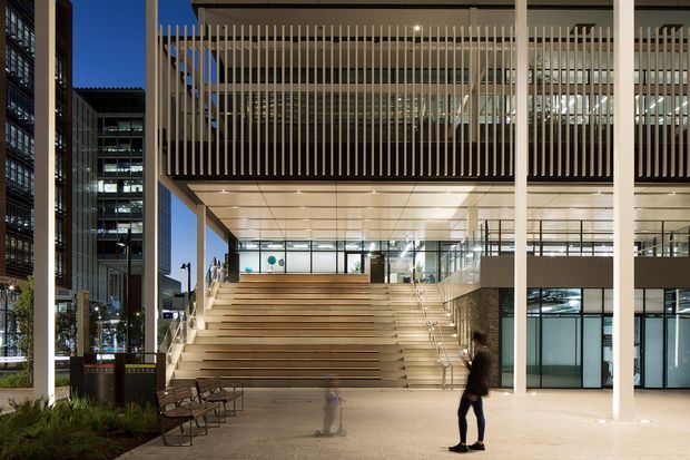 A contemporary multi-storey commercial building entrance at dusk, featuring a broad stepped public stair, glazed ground-level interiors, tall slender structural columns, and an upper-level terrace screened by closely spaced vertical fins. Adjacent office buildings and landscaped planting frame the urban plaza.