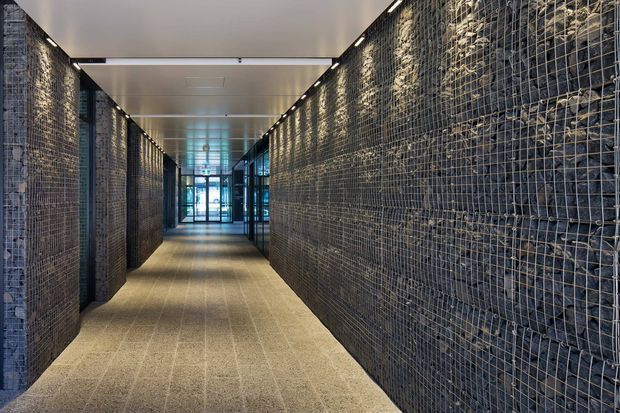 A long, modern interior corridor lined on both sides with exposed wire-mesh gabion walls filled with dark stone. The corridor has a pale carpeted floor, a smooth white ceiling with recessed linear lighting, and glazed doors and partitions at the far end. The strong repetition of the mesh and stone creates an industrial, textured architectural character.