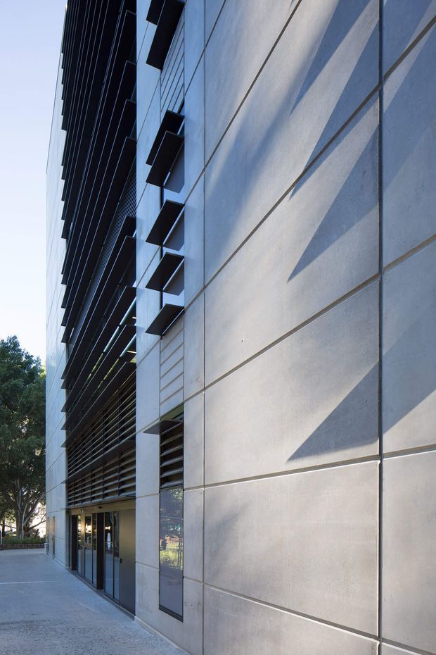 Exterior architectural photograph of a contemporary multi-storey building, viewed upward along a paved pedestrian edge. The facade combines large smooth grey concrete or cement panels with dark horizontal louvres, projecting box-like sunshades, narrow glazing, and recessed ground-level doors. Strong afternoon light creates angular shadows across the panelled facade, with trees visible at the edge of the site.