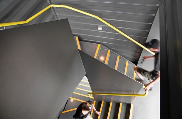 A dramatic overhead view looking down a modern industrial stairwell. Dark grey metal stairs, landings, walls and ceiling are accented by vivid yellow stair nosings and handrails. Several people are walking on the stairs, emphasizing the multi-level circulation space and angular geometry.