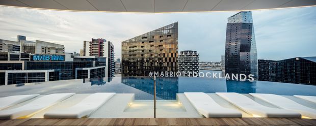 Wide rooftop terrace or poolside view overlooking a dense modern urban skyline, including prominent hotel and office towers. The foreground features a long reflective pool or water feature with white cushioned built-in loungers, framed beneath a curved architectural canopy. The scene has a polished contemporary hospitality feel.