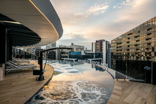 Rooftop hotel pool and terrace overlooking a dense urban docklands precinct. The scene features a curved architectural canopy with integrated linear lighting, timber decking, lounge chairs, a large infinity-edge pool with sculptural submerged forms, and surrounding multi-storey commercial and residential buildings.
