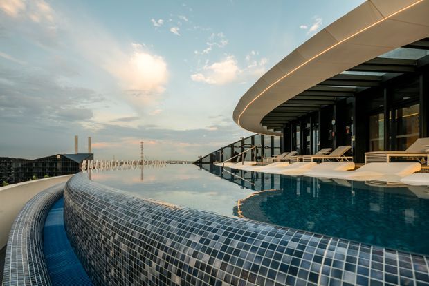 A rooftop infinity pool with a curved mosaic-tiled edge, surrounded by white loungers and a contemporary curved architectural canopy. The pool overlooks a distant urban skyline and is shown in warm evening light.