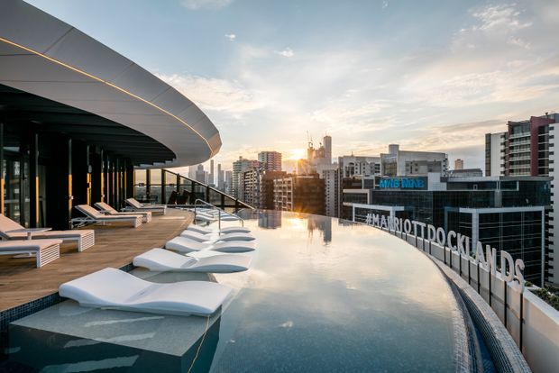 Rooftop infinity pool with white sculptural loungers on a timber deck, beneath a curved contemporary canopy. The pool overlooks a dense urban skyline at sunset, with a Marriott Docklands hotel sign visible on a neighbouring building.