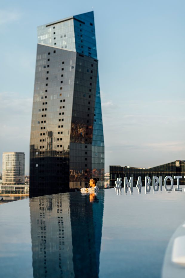 A rooftop infinity pool overlooking a modern urban skyline, dominated by a tall reflective glass-clad tower and adjacent commercial buildings. Large white “#MARRIOTT” lettering lines the pool edge, with a swimmer visible in the water. The scene is photographed at dusk with strong reflections across the calm pool surface.