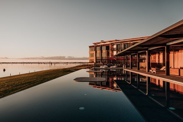 A modern waterfront building viewed at dusk, with extensive timber-clad structures, deep roof overhangs, balconies, and a long reflecting swimming pool in the foreground. Calm water and a low, open landscape extend beside the building, while warm interior lighting creates a dramatic reflection across the pool.