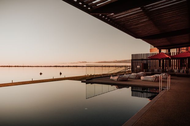 A tranquil waterfront outdoor terrace at dusk, featuring a dark reflective swimming pool in the foreground, timber pergola overhead, slatted screening, striped outdoor lounge seating, and red umbrellas overlooking calm water.
